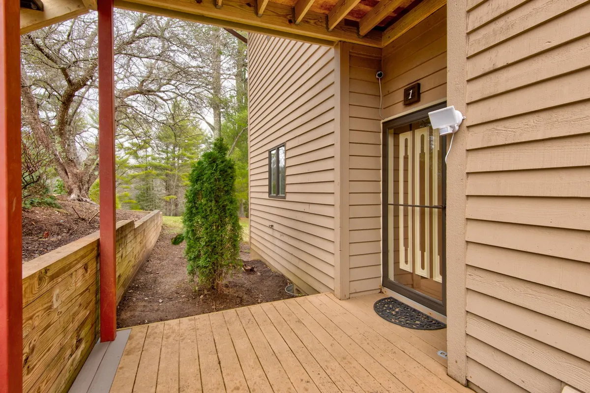 This exterior image features the front door of the condo unit, a welcoming wooden deck, and lush surrounding greenery. The scene suggests a space where families can gather outdoors, greet friends, and enjoy fresh air just steps from their home base.