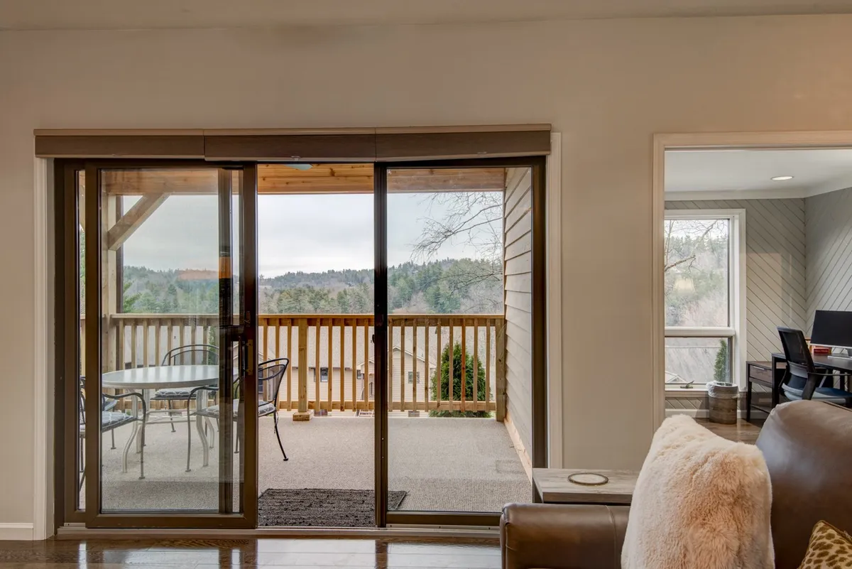 The image displays sliding glass doors opening onto a cozy patio with outdoor seating, offering a serene view of the surrounding landscape. Natural light streams into the interior, creating a seamless connection between the indoor living space and the private outdoor patio area.