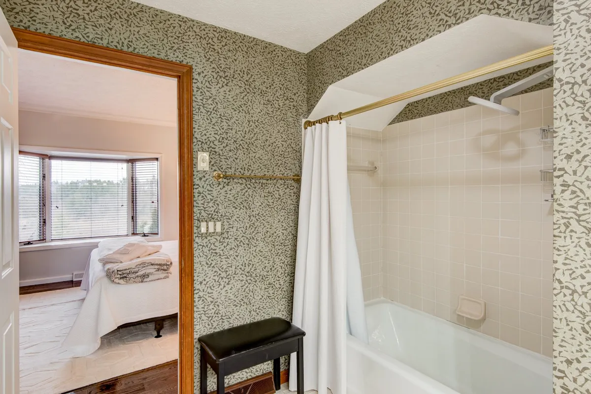 A bathroom featuring a relaxing bathtub with a wall pattern, a window offering a view, and a bed with fresh linens visible, blending bathing and sleeping spaces. The mood is calm, emphasizing relaxation and the scent of fresh linens.