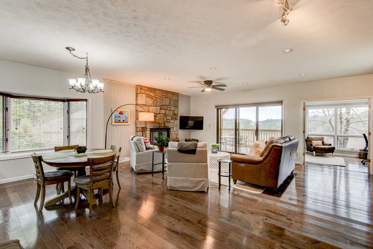 A bright, inviting dining and lounging space anchored by a stone fireplace. The area is furnished with a dining table and chairs, comfortable seating, and large windows that offer glimpses of the outdoors. The scene is perfect for hosting meals or enjoying the warmth of the fire after a day of exploration.