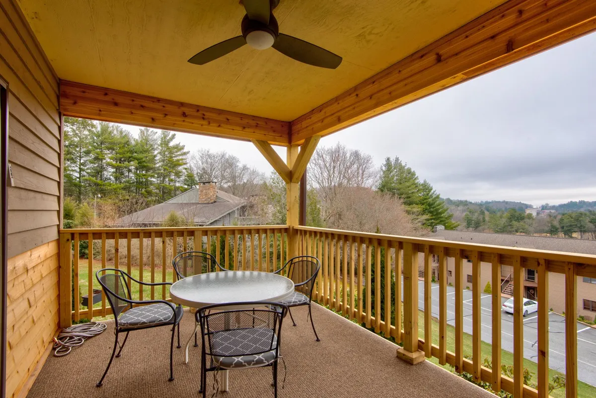 An exterior view of a balcony or patio featuring an outdoor dining table surrounded by lush trees and a sturdy railing. The scene is set for relaxed outdoor meals, gatherings, or quiet coffee moments, with the natural surroundings enhancing the sense of retreat.
