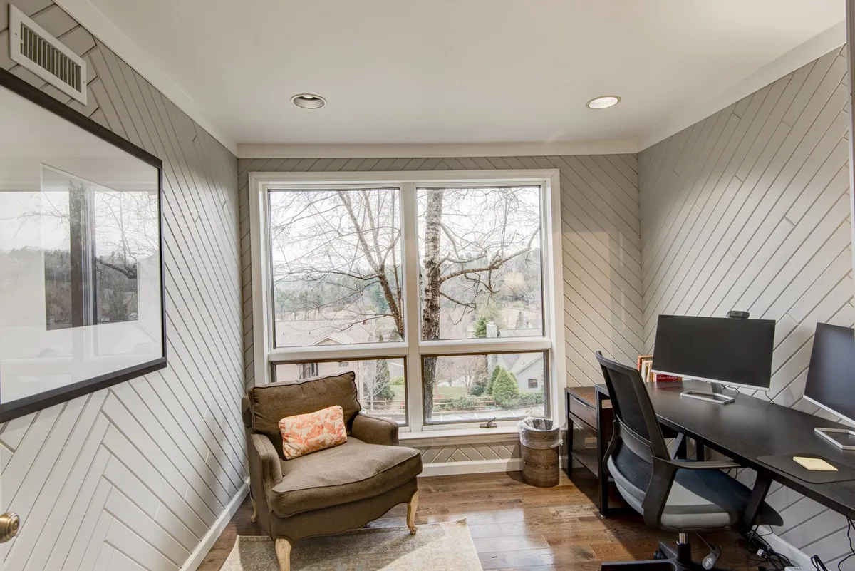 A modern sunroom office setup featuring a large desk, comfortable chair, and dual monitors positioned in front of expansive windows that provide abundant natural light and tranquil views of the outdoors. Subtle decorative touches add a calming ambiance, making the space ideal for focused work or creative pursuits.