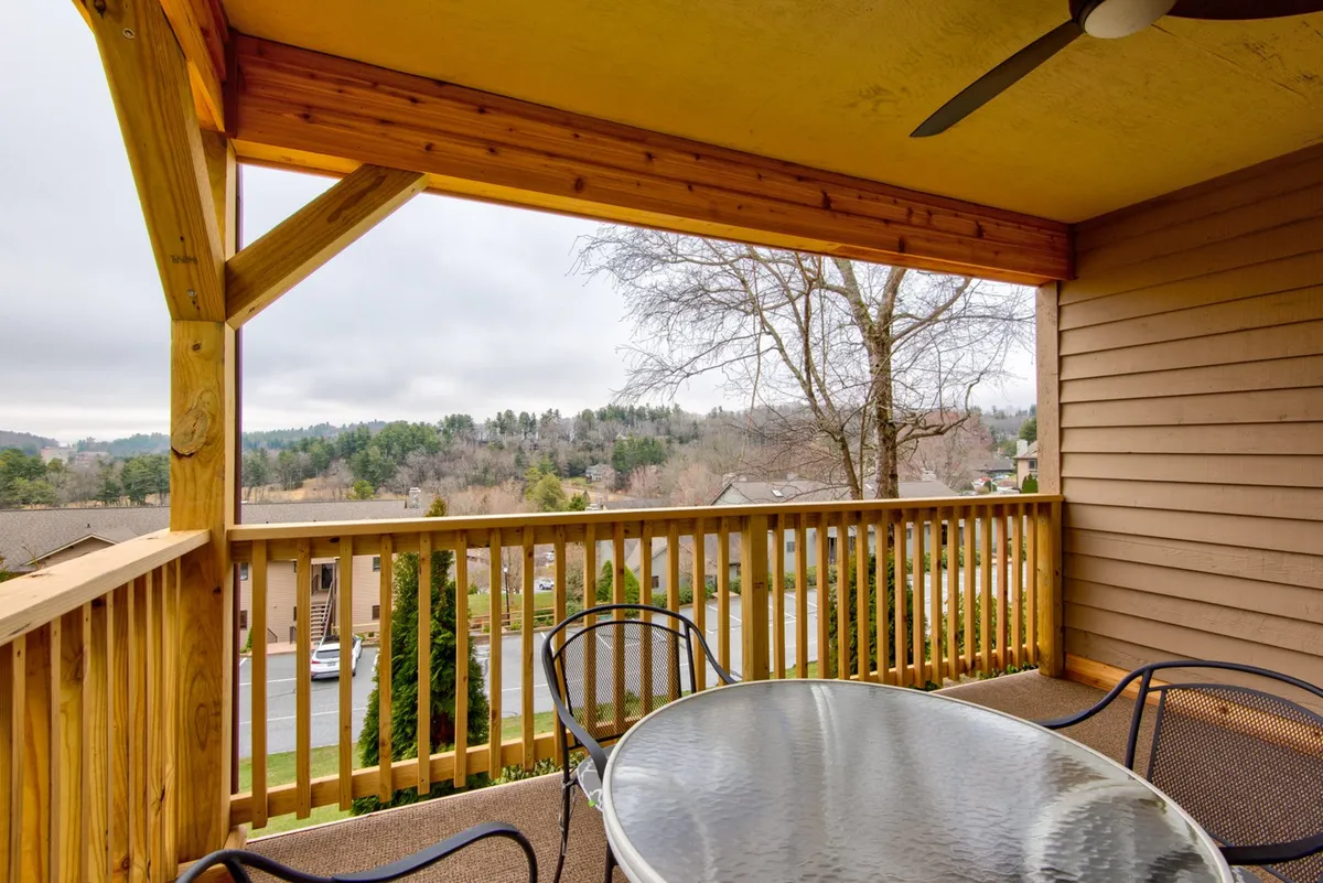A peaceful outdoor seating area with a wooden railing, glass table, and comfortable chairs overlooks trees and rolling hills. The space invites relaxation, socializing, and enjoying the scenic environment together as a family.