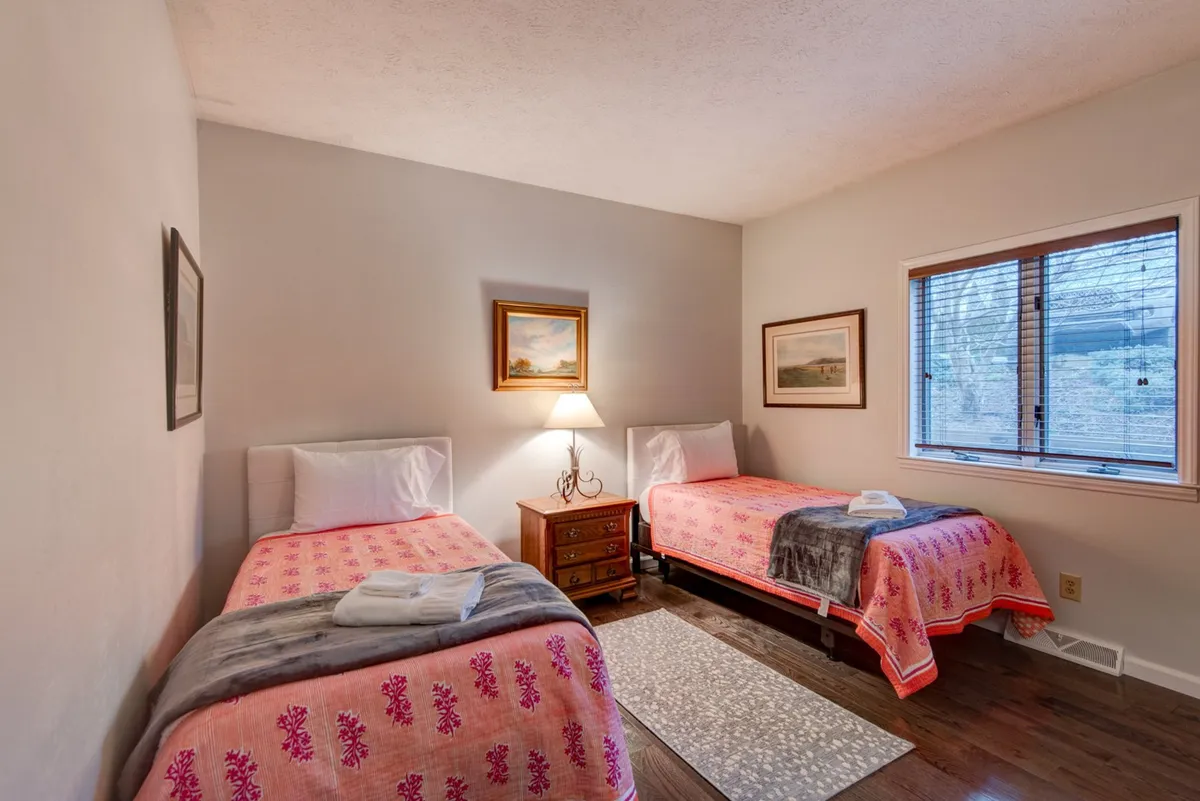 A bright and airy bedroom with two twin beds dressed in colorful bedding, a nightstand and lamp between them, and artwork on the walls. Light streams in through the window, creating a comfortable and relaxed vibe.