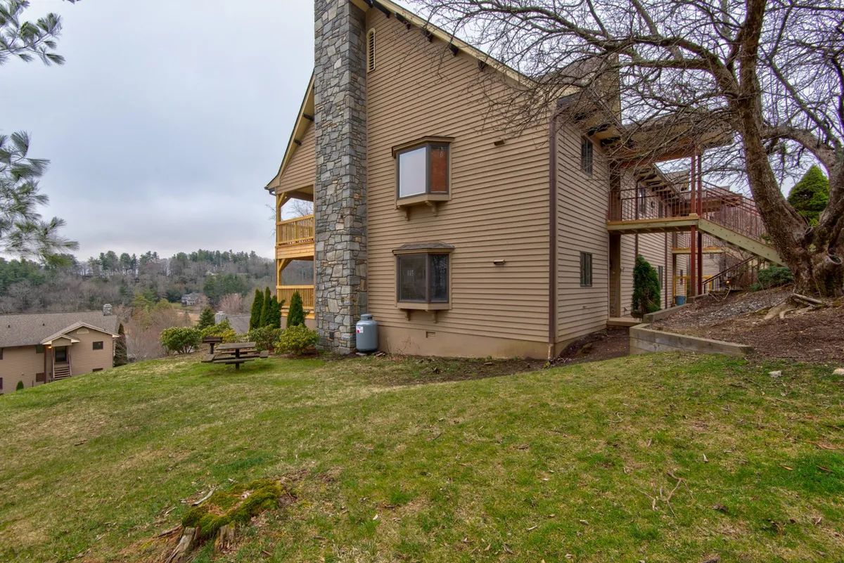 An exterior view of a building featuring a stone chimney, large windows that offer a glimpse of the inviting interior, wooden stairs, and a lush green lawn. The space feels private yet open to nature, ideal for outdoor gatherings or enjoying quiet moments outside.