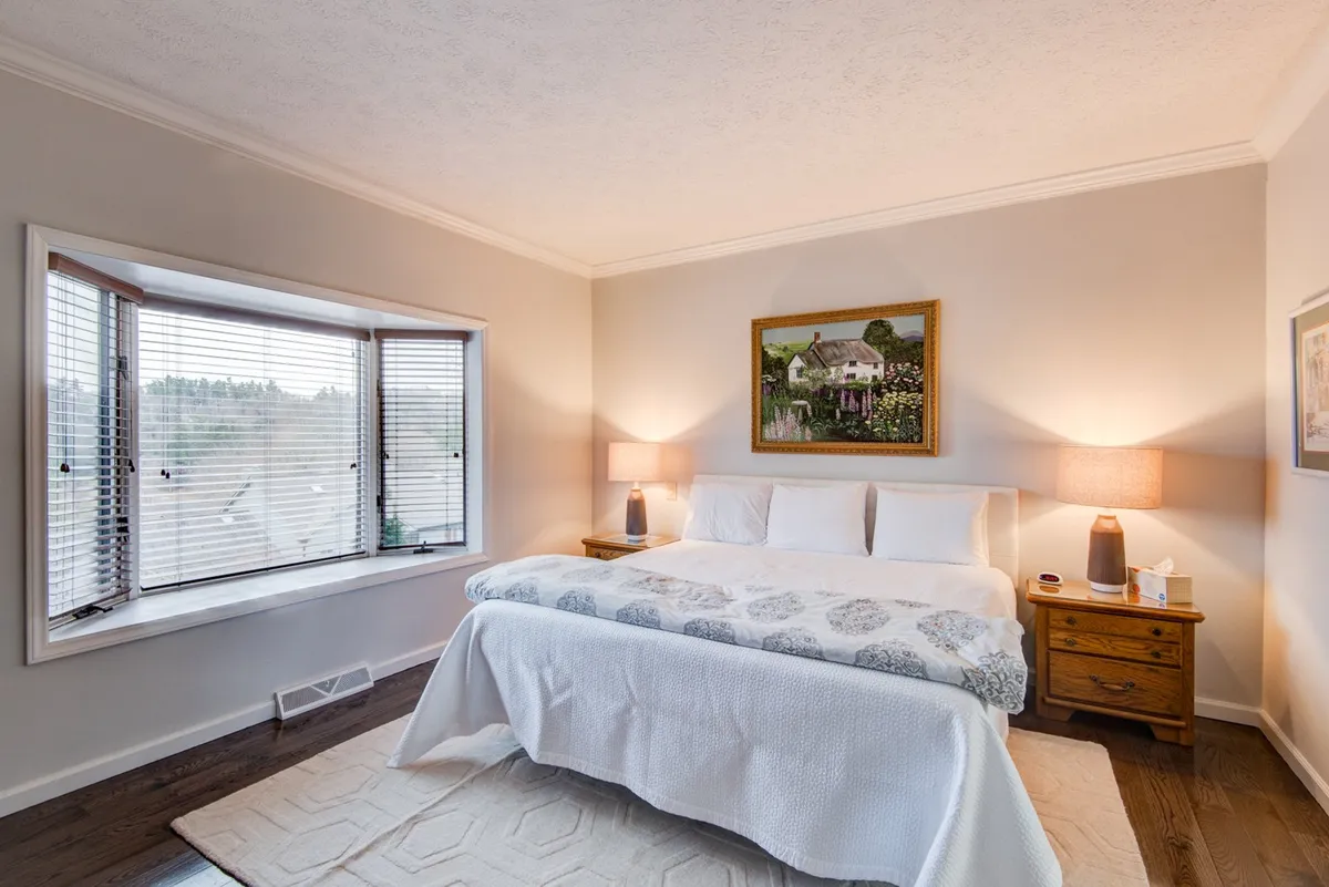 A tranquil bedroom with a neatly made bed featuring crisp white linens, a framed painting above the headboard, and a window with blinds letting in gentle natural light. The atmosphere is peaceful, inviting relaxation and rest.